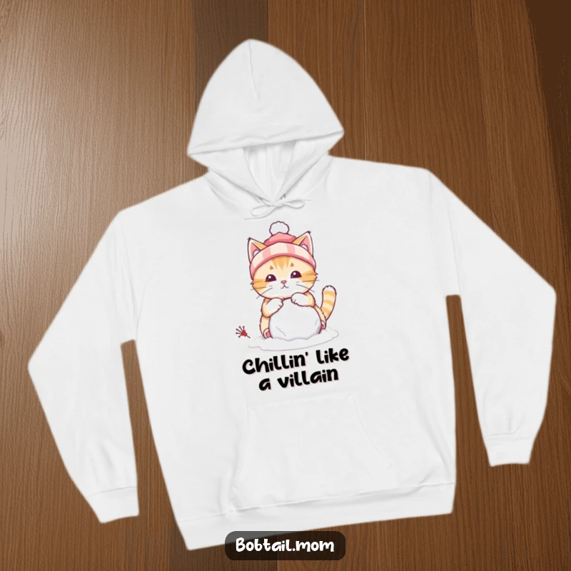 Funny hoodie showing a bobtail cat wearing a winter hat, focused on constructing a small snowball, embodying winter playfulness.