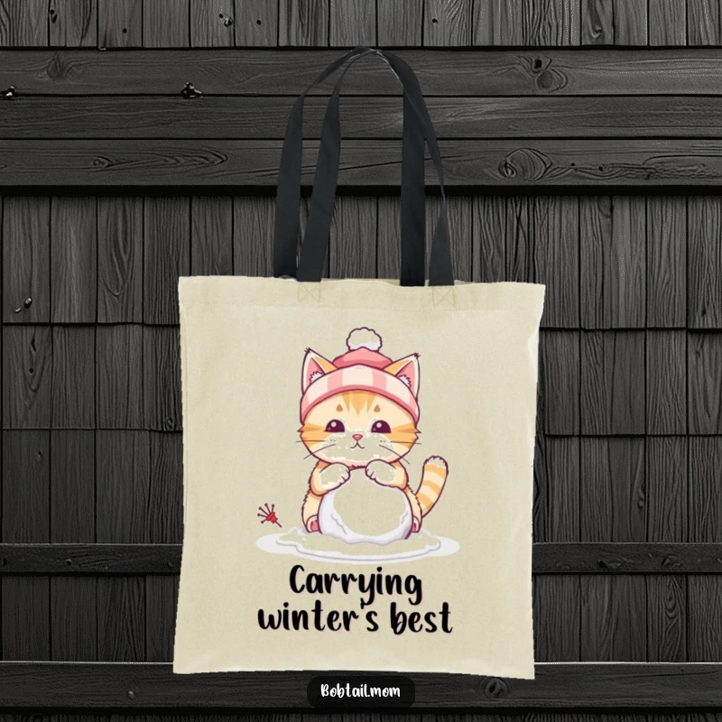 Funny tote bag featuring a bobtail cat with a winter hat, building a tiny snowball, ready for snowy outings.